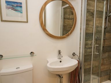 A bathroom with a round wooden mirror above a white sink and a glass shower with tiled walls at Tan y Craig in Solva