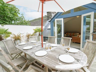 An outdoor wooden dining table with plates glasses and cutlery under a large umbrella on a patio with garden plants and open doors to a living room at Tan y Craig in Solva