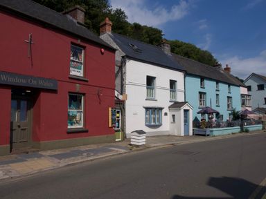 A street with red white and blue buildings including a shop and outdoor seating with umbrellas at Tan y Craig in Solva