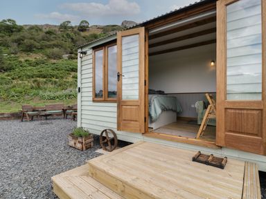 The open wooden door of a cabin showing a bed inside and outdoor seating on gravel in a rural area at Tan Y Castell Shepherds Hut in Llangollen