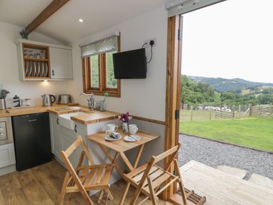 A small kitchen with wooden countertops a sink and a table with two chairs next to an open door showing a green landscape at Tan Y Castell Shepherds Hut in Llangollen