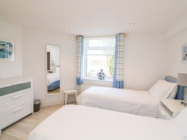 A bedroom with two single beds a window with blue and white curtains a chest of drawers and a mirror at 1 Camperdown Lodge in Salcombe