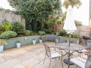 An outdoor patio area with a round metal table and four chairs with striped cushions a closed umbrella and potted plants along a wooden raised garden bed at 1 Camperdown Lodge in Salcombe