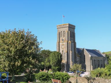 A stone church with a clock tower and a flagpole surrounded by trees and benches at 1 Camperdown Lodge in Salcombe