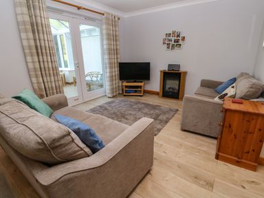 A living room with two beige sofas a television on a stand a small electric fireplace a wooden drawer unit and glass doors with curtains leading to a sunroom at Primrose Cottage Goldenbank near Falmouth