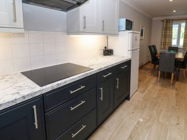 A kitchen with black lower cabinets white upper cabinets a marble countertop an electric stovetop a white refrigerator with a microwave on top and a dining area with a wooden table and chairs at Primrose Cottage in Goldenbank near Falmouth