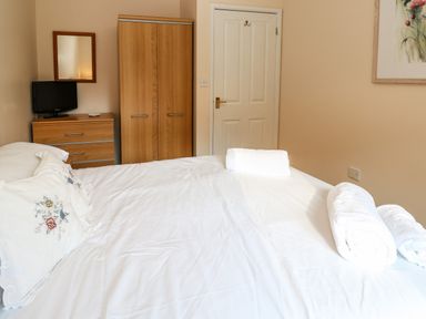 A bedroom with a bed with white sheets and pillows a wooden wardrobe a chest of drawers with a small television a door and a framed picture on the wall at Primrose Cottage in Goldenbank near Falmouth