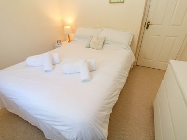A bedroom with a double bed with white bedding and rolled towels a bedside table with a lamp and clock a chest of drawers and a closed door at Primrose Cottage in Goldenbank near Falmouth