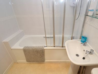 A bathroom with a bathtub featuring a folding glass shower screen a sink with soap and a gray towel on the bathtub edge at Primrose Cottage Goldenbank near Falmouth