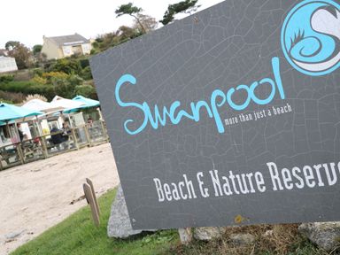 A weathered signboard for Swanpool Beach and Nature Reserve near a fenced seating area with blue umbrellas and houses on a hillside in the background at Primrose Cottage in Goldenbank near Falmouth