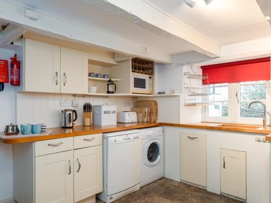 A kitchen with white cabinets wooden countertops a dishwasher a washing machine a microwave a kettle a toaster a bread box and a window with a red blind at St Samson in Port Isaac
