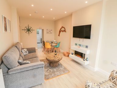 A living room with a gray sofa a round coffee table a wall-mounted tv a dining table with colorful chairs and decorations at Bay View in Criccieth