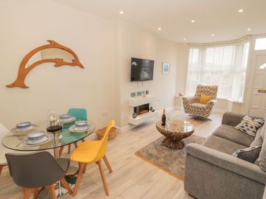 A living room with a round glass dining table with multicolored chairs a grey sofa patterned armchair a coffee table a wall-mounted TV and a dolphin-shaped mirror at Bay View in Criccieth