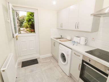 A kitchen with white cabinets a washing machine an oven a countertop with kettle toaster and cups a sink and an open door showing outdoor plants at Bay View in Criccieth