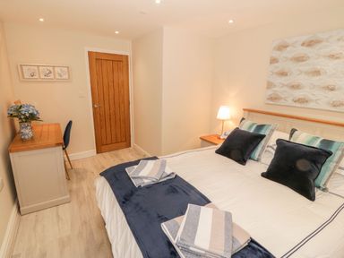 A bedroom with a bed, pillows, a bedside table with a lamp, a wooden door, a dresser with a vase of flowers, and a chair at Bay View in Criccieth
