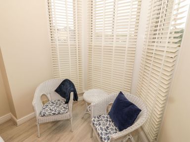 A corner seating area with two white wicker chairs and a matching small round table with window blinds behind at Bay View in Criccieth