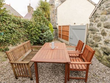 An outdoor patio area with wooden table and chairs on gravel near a stone wall and a garden with bushes and a wooden gate