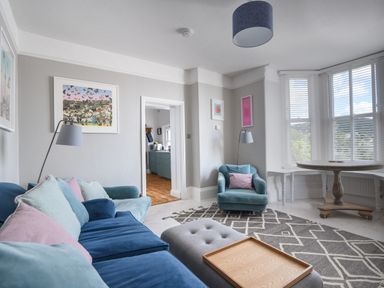 A living room with a blue sofa a blue armchair a gray ottoman wooden tray on ottoman round wooden table and large windows with blinds at Coastal View in Lyme Regis