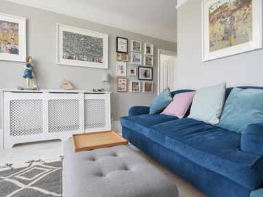 A living room with a blue sofa and pastel cushions a grey ottoman with a wooden tray a white radiator cover and framed pictures on walls at Coastal View in Lyme Regis