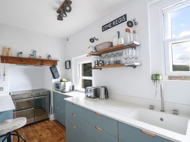 A kitchen with blue cabinets a white countertop and wooden shelves with jars and glasses at Coastal View in Lyme Regis