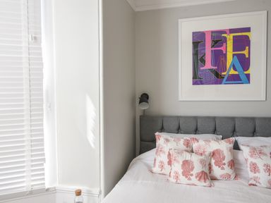A bedroom with a grey upholstered headboard white bedding with red patterned pillows a framed colorful abstract artwork on the wall and white window blinds