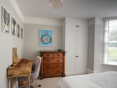 A bedroom with a wooden desk and chair wooden chest of drawers white wardrobe and window with blinds at Coastal View in Lyme Regis
