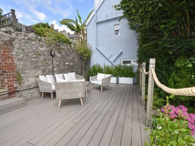 An outdoor patio with wicker chairs and cushions next to a stone wall and a blue house at Coastal View in Lyme Regis