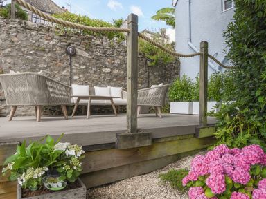 An outdoor seating area with sofas cushions and a coffee table on a wooden deck with flowers and plants surrounding at Coastal View in Lyme Regis