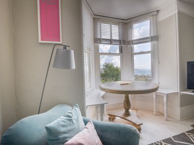 A corner with a round wooden table near bay windows a blue armchair with cushions and a floor lamp at Coastal View in Lyme Regis
