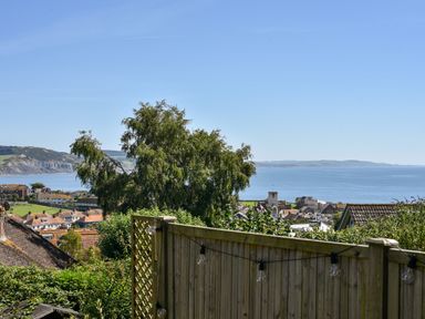 A view of rooftops trees and the sea beyond a wooden fence with string lights at Coastal View in Lyme Regis