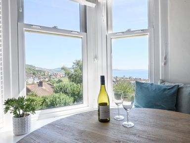 A round wooden table with a bottle of wine and two wine glasses next to a bay window with a view of houses trees hills and the sea at Coastal View in Lyme Regis