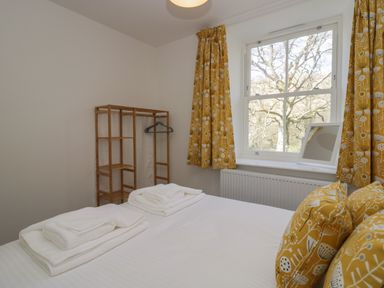 A bedroom with a bed, folded towels, wooden clothes rack, window with yellow patterned curtains and cushions at Dalegarth Hall Farm Cottage 1 Boot near Eskdale Green