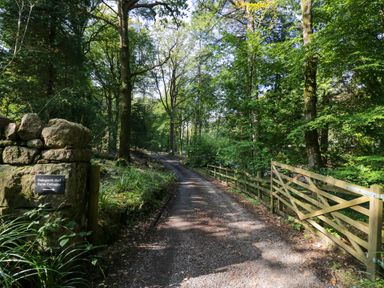 A gravel driveway surrounded by trees with a stone wall and sign for Dalegarth Hall Farm Cottages at Boot near Eskdale Green