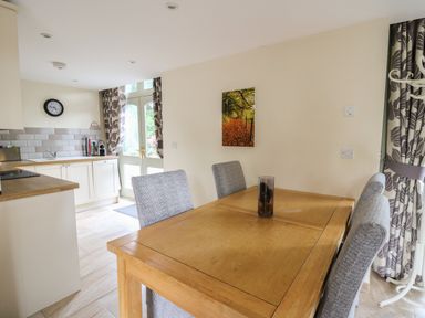 A dining area with a wooden table and four upholstered chairs near a kitchen with white cabinets and a door with patterned curtains at Gelli Barn Near Llandeilo