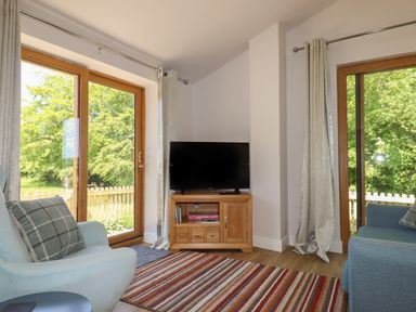 A living room with a TV on a wooden stand between two large windows at Adipit Stables Buckland Brewer near Bideford
