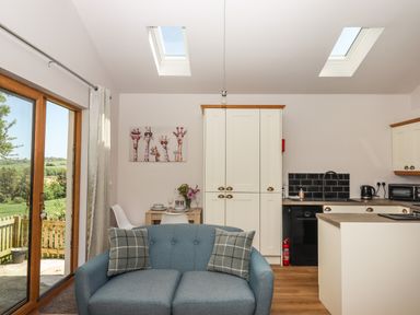 A living area with a blue sofa and two checkered pillows next to a small dining table with two chairs and a kitchen with white cabinets and black appliances at Adipit Stables in Buckland Brewer near Bideford