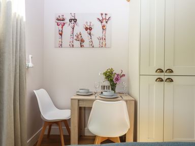 A small dining area with two white chairs a wooden table set with plates bowls glasses and a flower vase a giraffe painting on the wall and cream cabinets nearby at Adipit Stables Buckland Brewer near Bideford