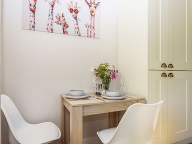 A small dining area with a wooden table set for two with bowls plates and glasses two white chairs flowers in a vase and a colorful giraffe artwork on the wall at Adipit Stables Buckland Brewer near Bideford