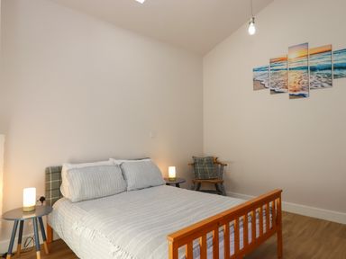 A bedroom with a wooden bed a chair with a cushion two side tables with lamps and a multi-panel beach painting on the wall at Adipit Stables Buckland Brewer near Bideford