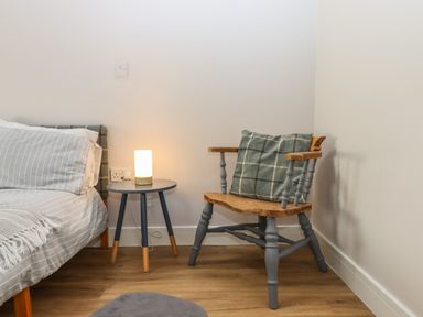 A bedroom corner with a bed with striped bedding a round side table with a lamp and a wooden chair with a cushion at Adipit Stables in Buckland Brewer near Bideford