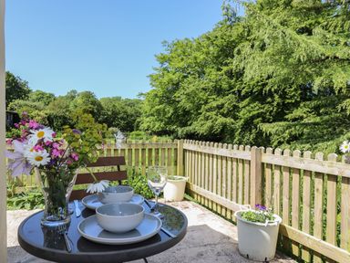 A patio table with dishes and a vase of flowers beside a wooden fence with potted plants and trees in the background at Adipit Stables Buckland Brewer near Bideford