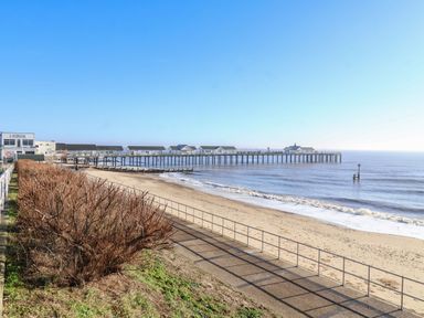 A sandy beach with a pier extending into the sea and a pathway with a railing alongside the beach at Pebbles in Southwold