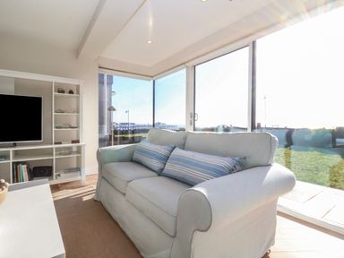 A living room with a white sofa and striped cushions facing a wall unit with a television and shelves at Pebbles in Southwold