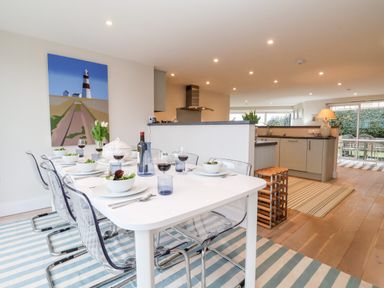 A dining area with a white table set with bowls and glasses and transparent chairs on a striped rug next to a kitchen with cabinets and a lamp at Pebbles Southwold