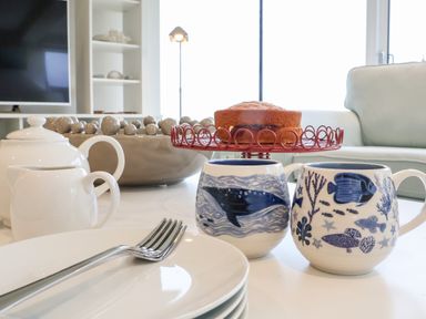 A table set with two blue and white mugs, a small cake on a red wire stand, a white teapot and creamer, and stacked white plates with a fork in a living room at Pebbles in Southwold