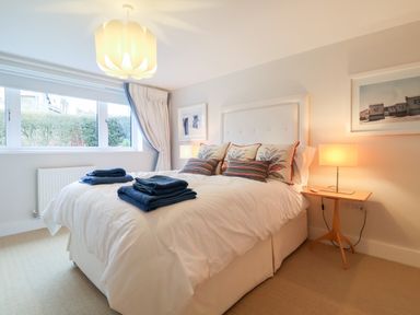 A bedroom with a double bed dressed in white linens and decorative pillows with folded towels on top next to a window with a curtain and a bedside table with a lamp at Pebbles in Southwold