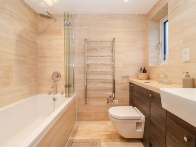 A bathroom with a bathtub shower combination a wall-mounted toilet wooden cabinets and a towel radiator at Pebbles in Southwold