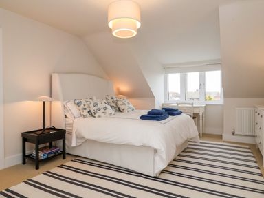 A bedroom with a white bed with patterned pillows a black side table with a lamp striped rug and a desk by the window at Pebbles in Southwold