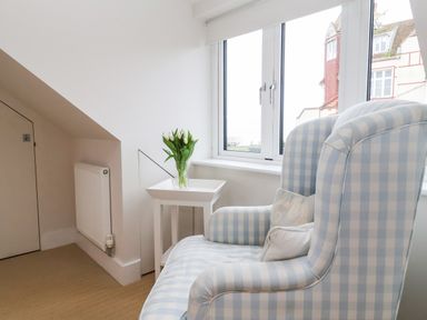 A corner with a blue and white checkered armchair next to a white side table with a vase of flowers by a window at Pebbles in Southwold