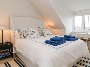 A bedroom with a bed with white and blue bedding and blue towels on it with nightstands and lamps and a window with a desk and chair at Pebbles Southwold